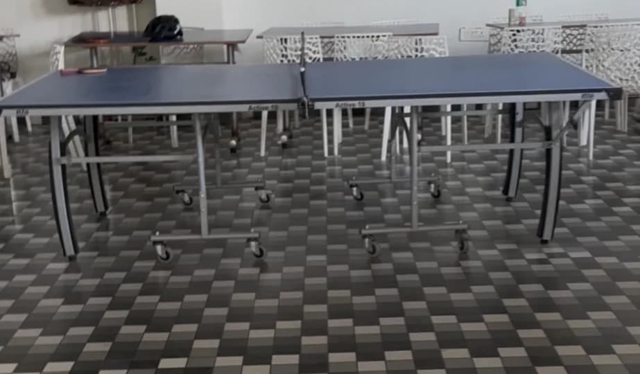 Table tennis table in the cafeteria, with tables and white lattice-pattern chairs behind it on a checkered tile floor