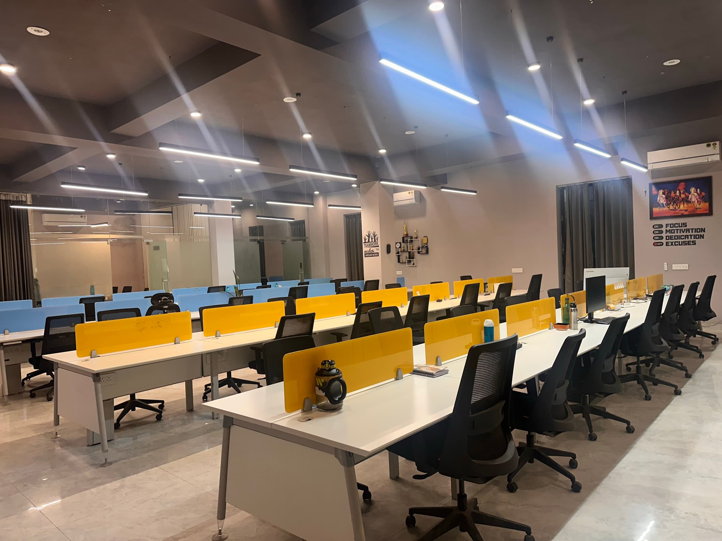 Rows of coworking desks with yellow dividers and mesh chairs, facing a wall graphic of switches: Focus, Motivation and Dedication on, Excuses off